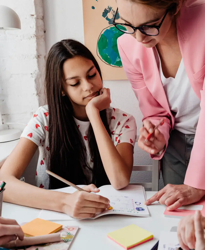 A young girl with a pen and notepad being taught by a lady.